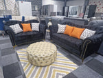 Load image into Gallery viewer, An elegant black Chesterfield sofa with button-tufted design, scroll arms, and metallic-studded detailing, accompanied by plush orange and white pillows, placed in a showroom with modern decor.
